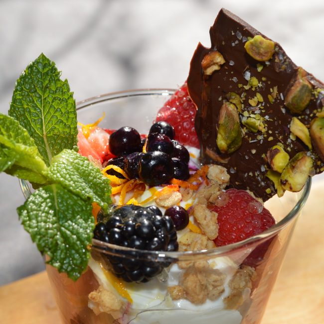 Close up photo of a fresh vanilla yogurt parfait with granola, berries, and pistachio-chocolate bark.